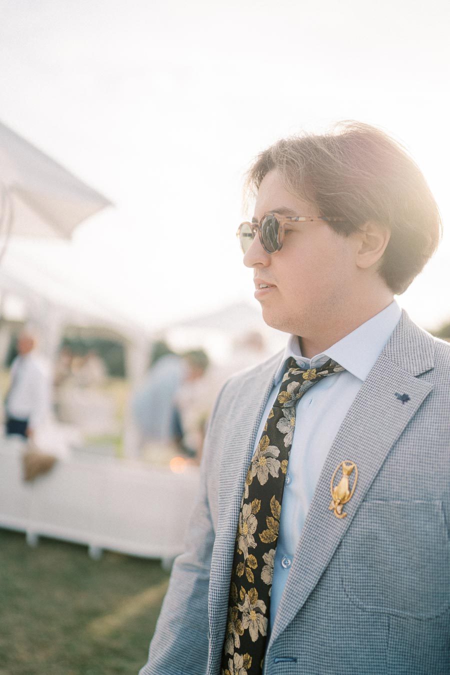 A man in sunglasses and a light gray suit with a floral tie at an outdoor event, with a soft, sunlit background.