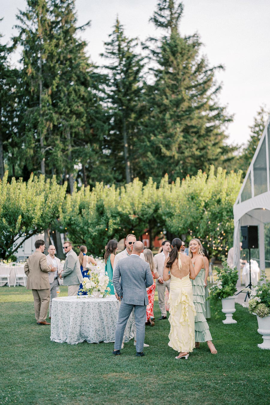Guests mingling at an outdoor wedding reception, elegantly dressed, surrounded by lush greenery and a beautifully decorated table.
