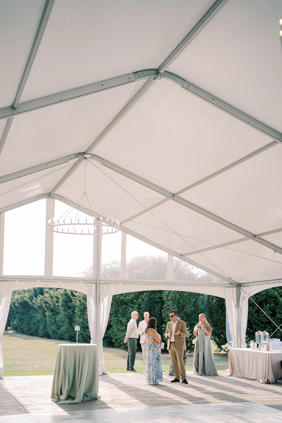 Elegant outdoor tented event with people socializing, surrounded by lush greenery, featuring a chandelier and tables with linens, creating a serene and sophisticated atmosphere.