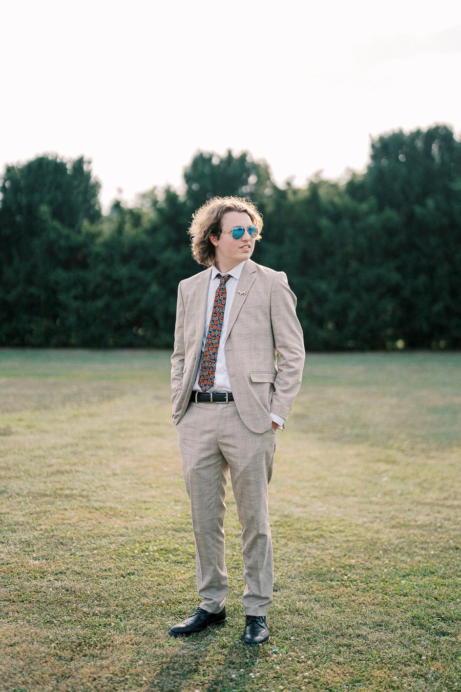 A person wearing a light beige suit with a colorful tie stands confidently on a grassy field, sporting blue sunglasses, with trees in the background.