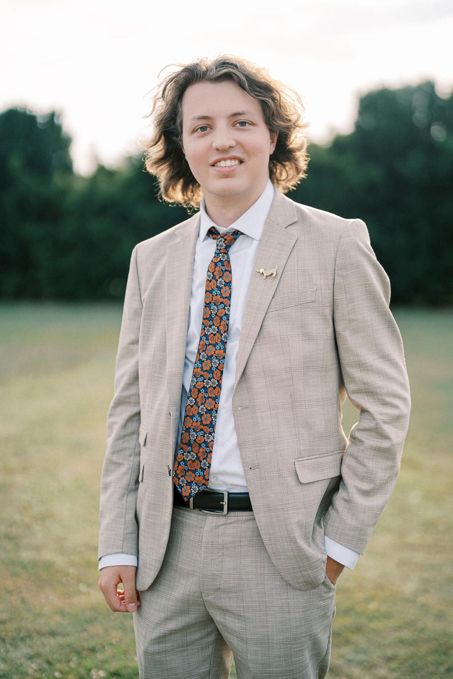 A young man with curly hair wearing a beige suit and patterned tie stands outdoors, smiling with hands in pockets, against a blurred green background.