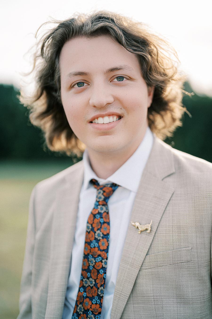 Smiling individual with curly hair wearing a beige suit and floral tie, standing outdoors on a sunny day.