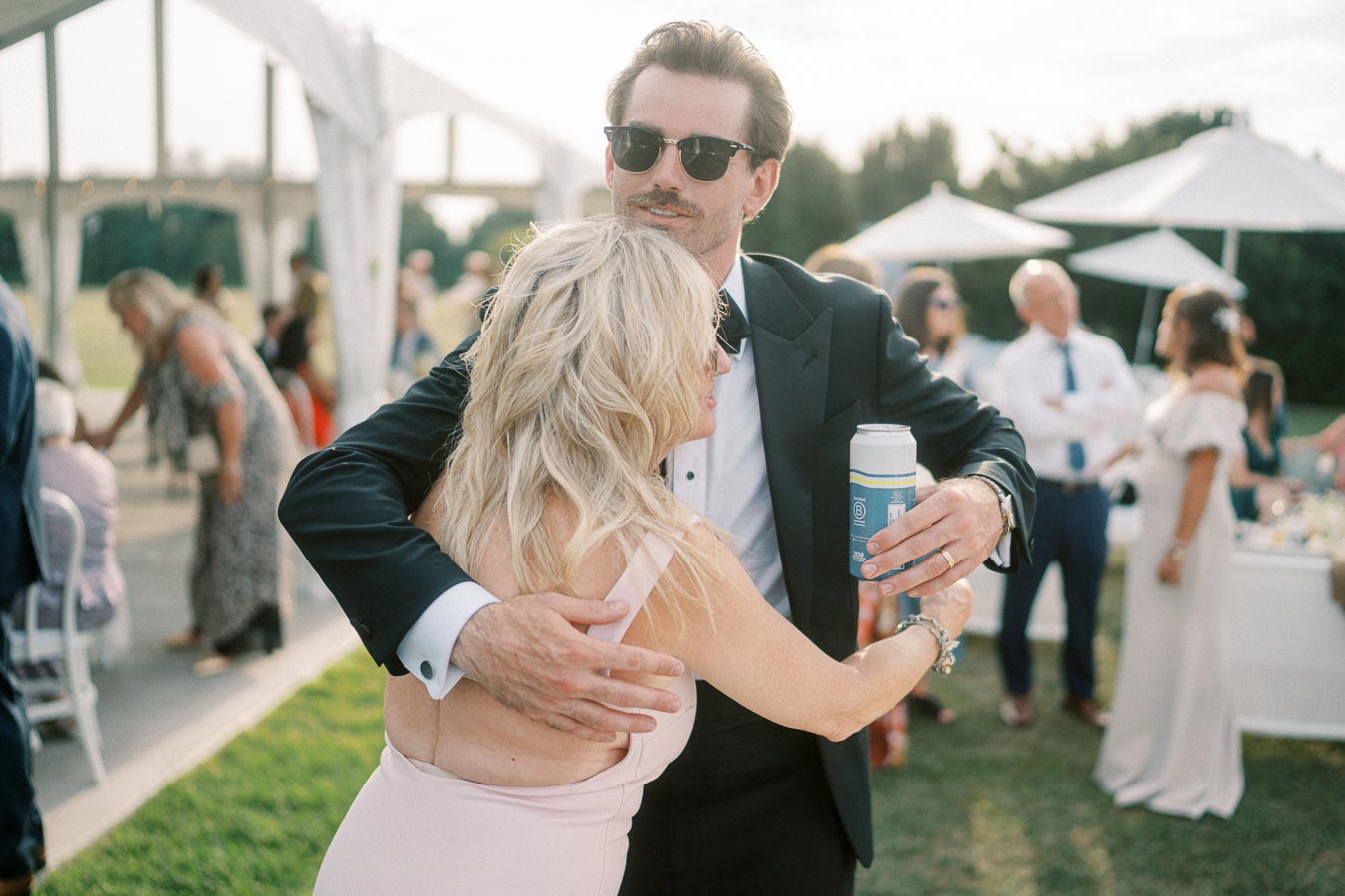 A man in sunglasses and a suit warmly embracing a woman in a pink dress at an outdoor event, surrounded by casually dressed people and umbrella tables.
