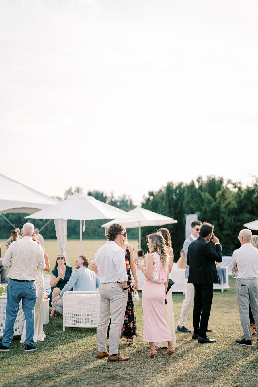A group of elegantly dressed people socializing outdoors at a garden party, with white umbrellas and greenery in the background.