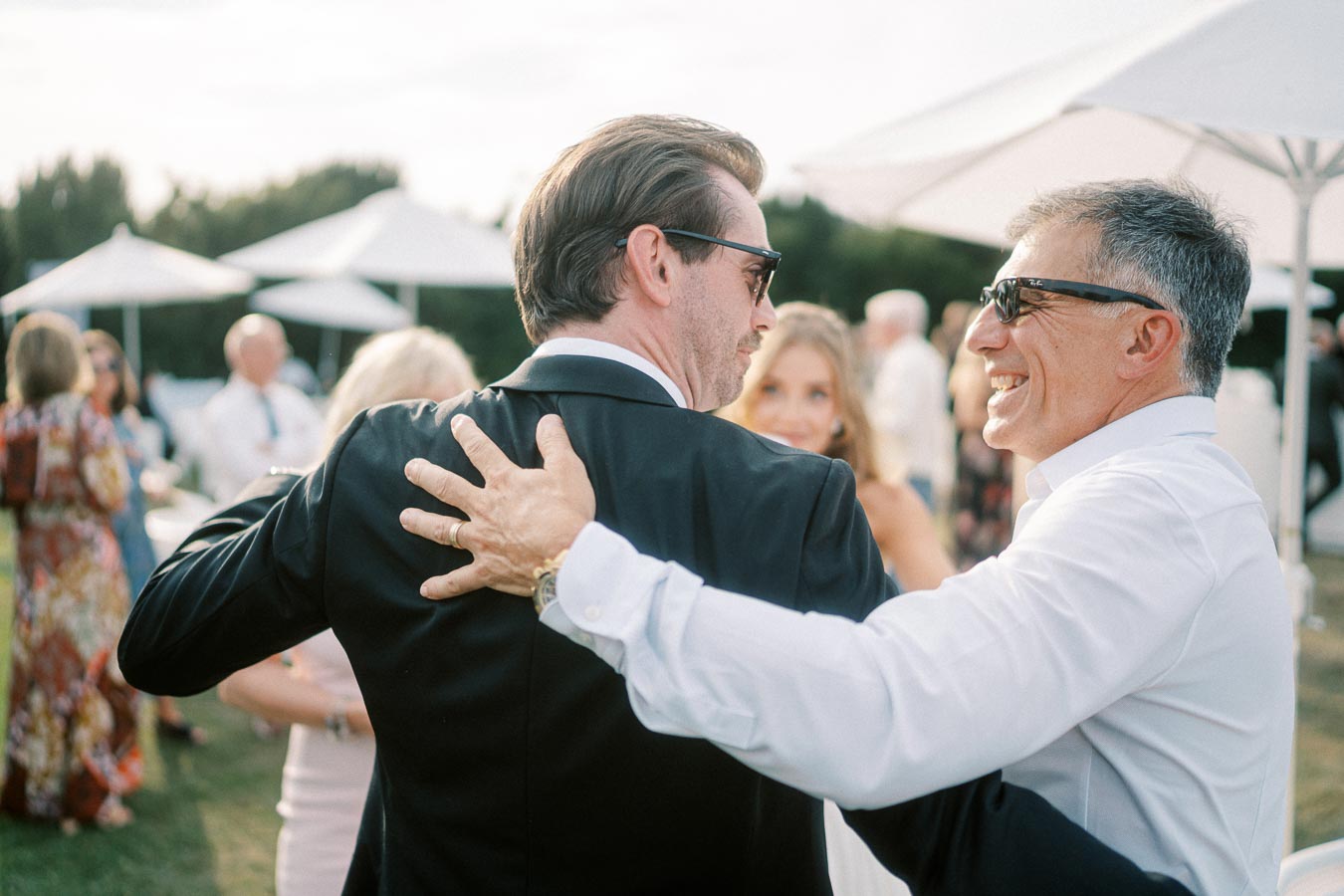 Two men in sunglasses share a joyful moment and embrace at an outdoor social event, with other smiling attendees and white umbrellas in the background, exuding a festive and friendly atmosphere.