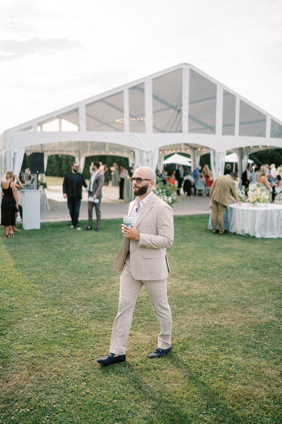 A man in a light beige suit walks across a grassy area with a drink in hand, with a white event tent and multiple people in the background, creating a festive outdoor gathering scene.