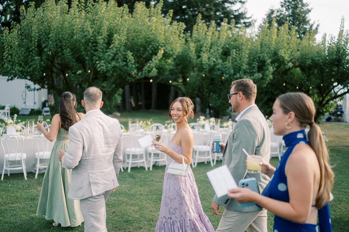 Group of elegantly dressed guests enjoying a garden wedding reception, with a woman in a lavender dress smiling at the camera, surrounded by lush greenery and beautifully arranged tables.