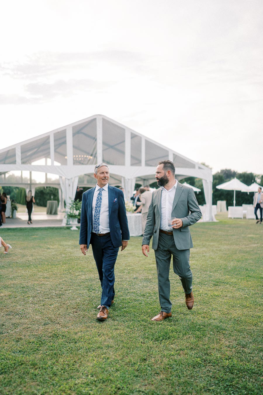 Two men in suits walking on a grassy lawn outside a white tented event venue, with a relaxed social atmosphere in the background.
