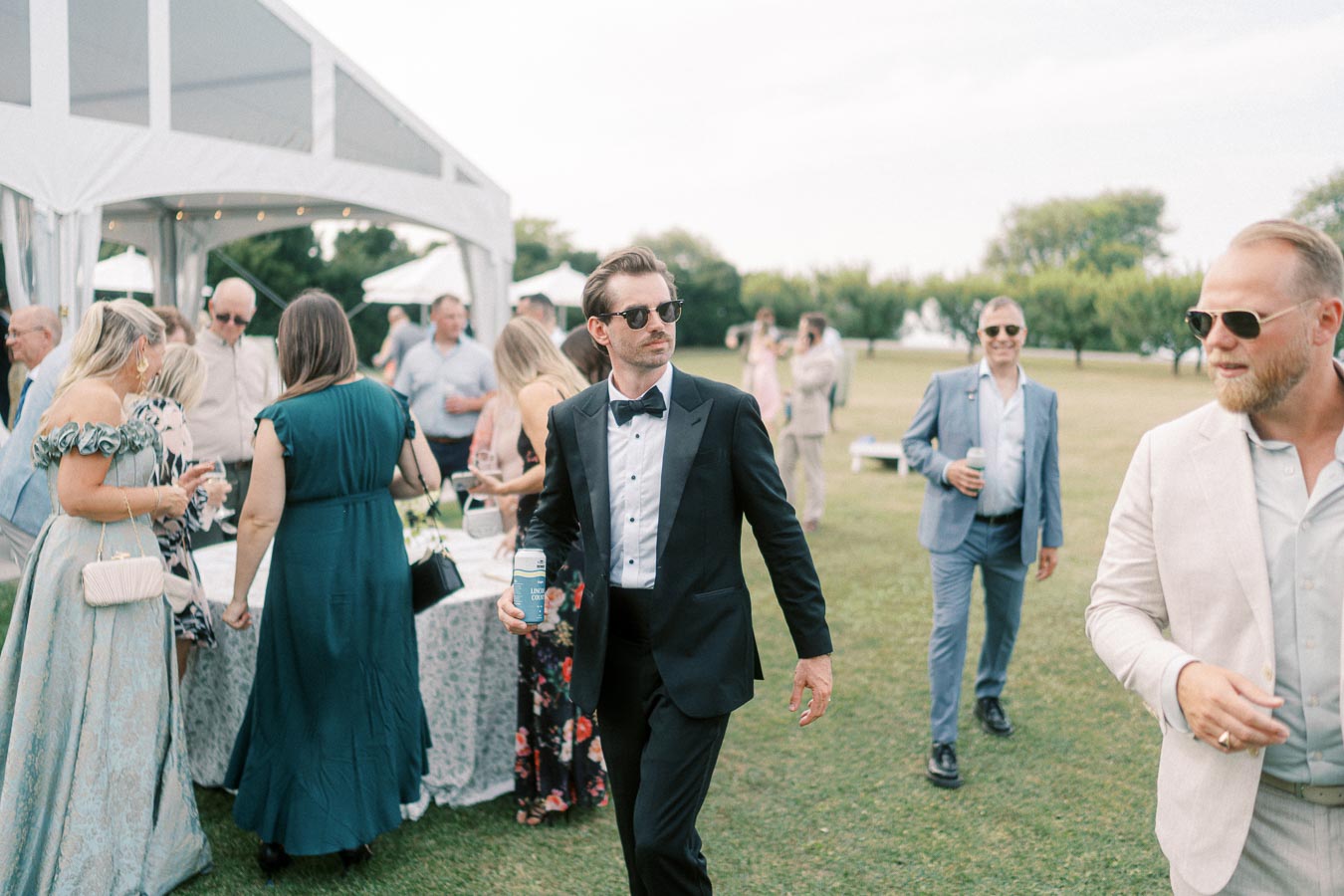 A stylish outdoor wedding reception with guests mingling; a man in a black tuxedo and sunglasses stands in the foreground, with elegantly dressed attendees and a large tent in the background.