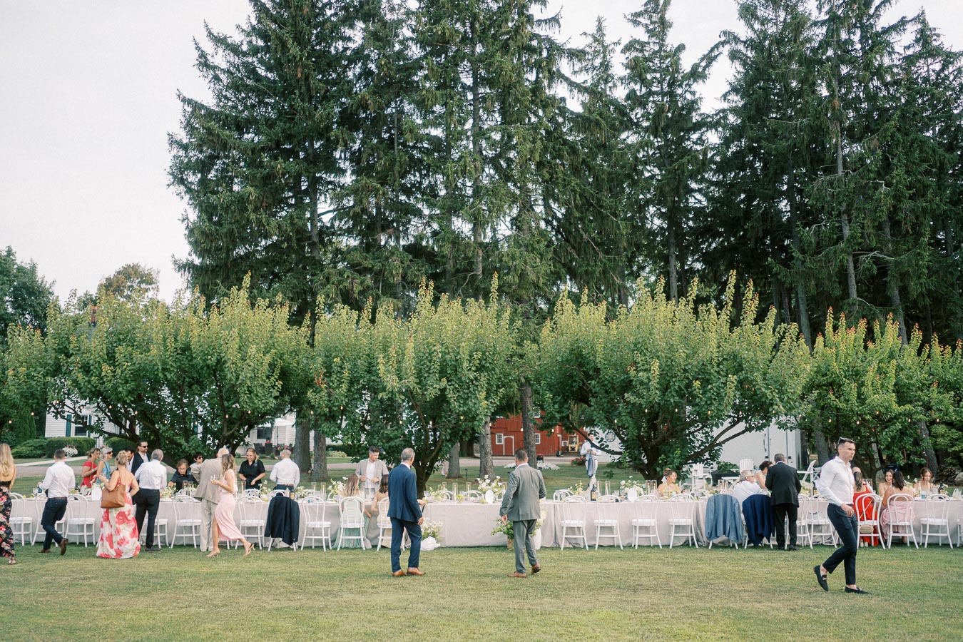 Outdoor wedding reception with guests mingling around elegantly set tables in a garden setting, surrounded by lush green trees and natural landscaping.