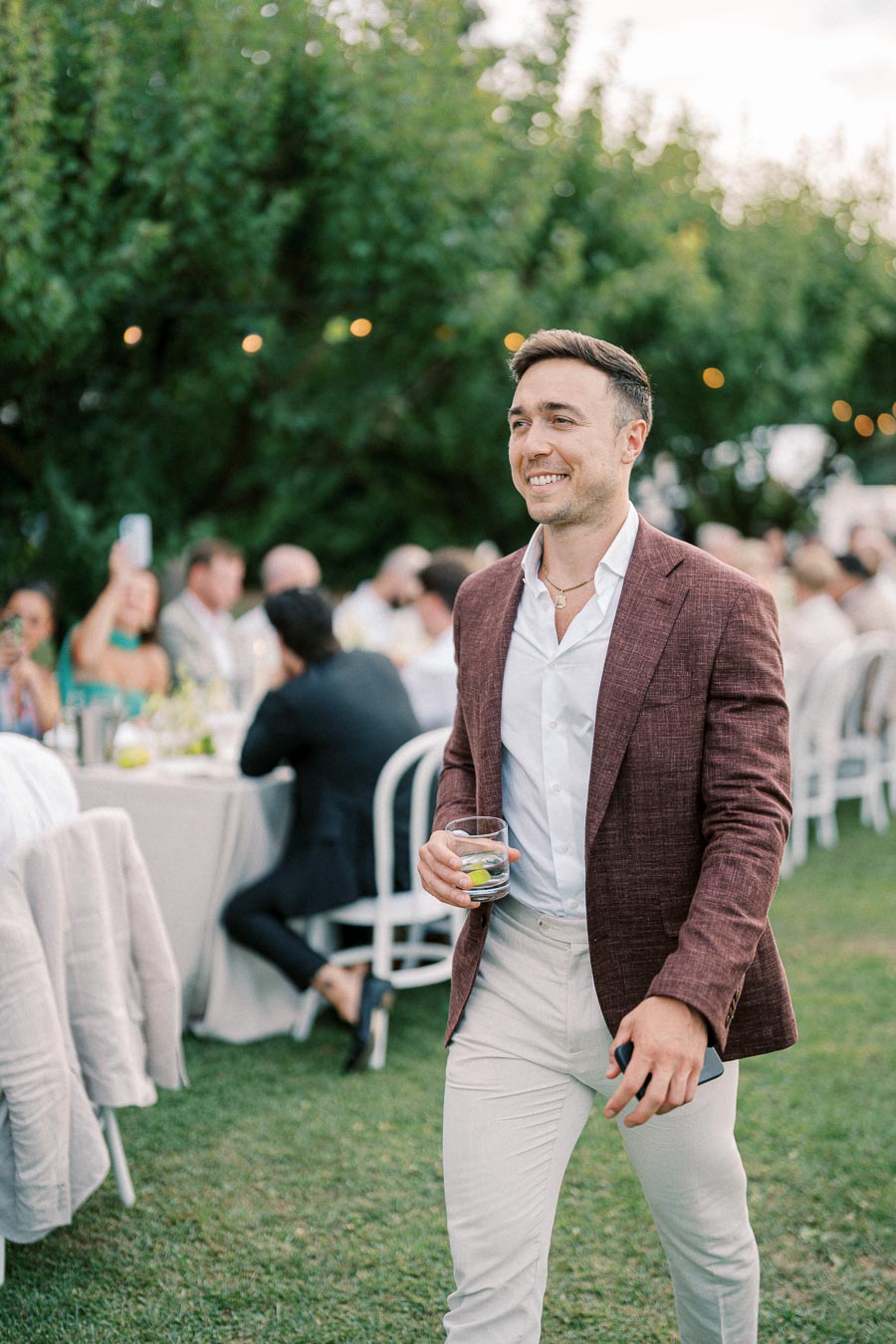 A man in a maroon blazer and white shirt is smiling while holding a drink and walking on grass at an outdoor event with people seated at tables in the background.