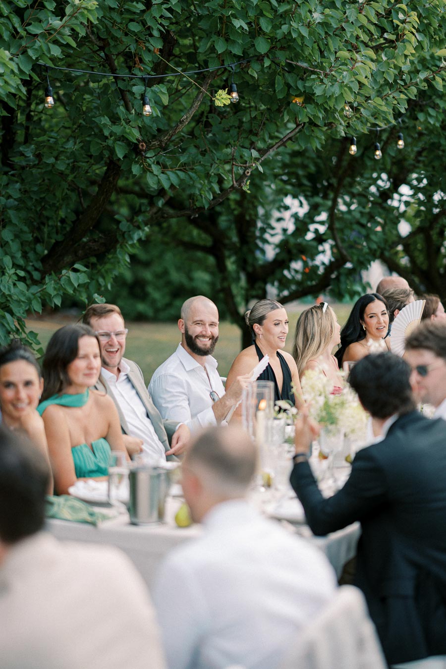 A group of people enjoying an outdoor dinner party under string lights, seated at a long table adorned with flowers and candles, surrounded by lush greenery.