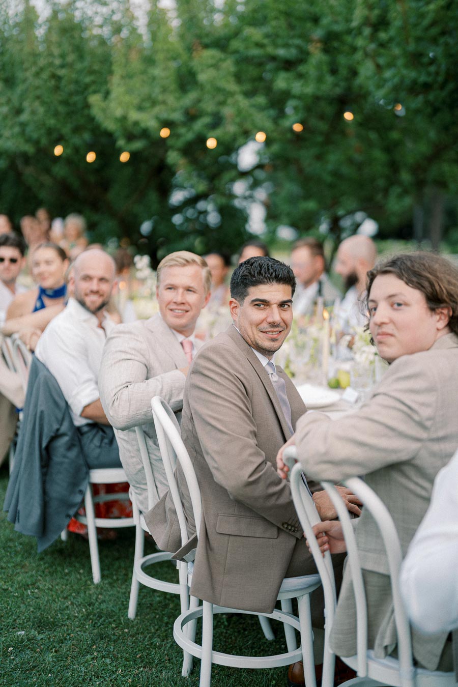 Guests seated outdoors at a wedding reception, dressed in light-colored suits, with greenery and string lights in the background.