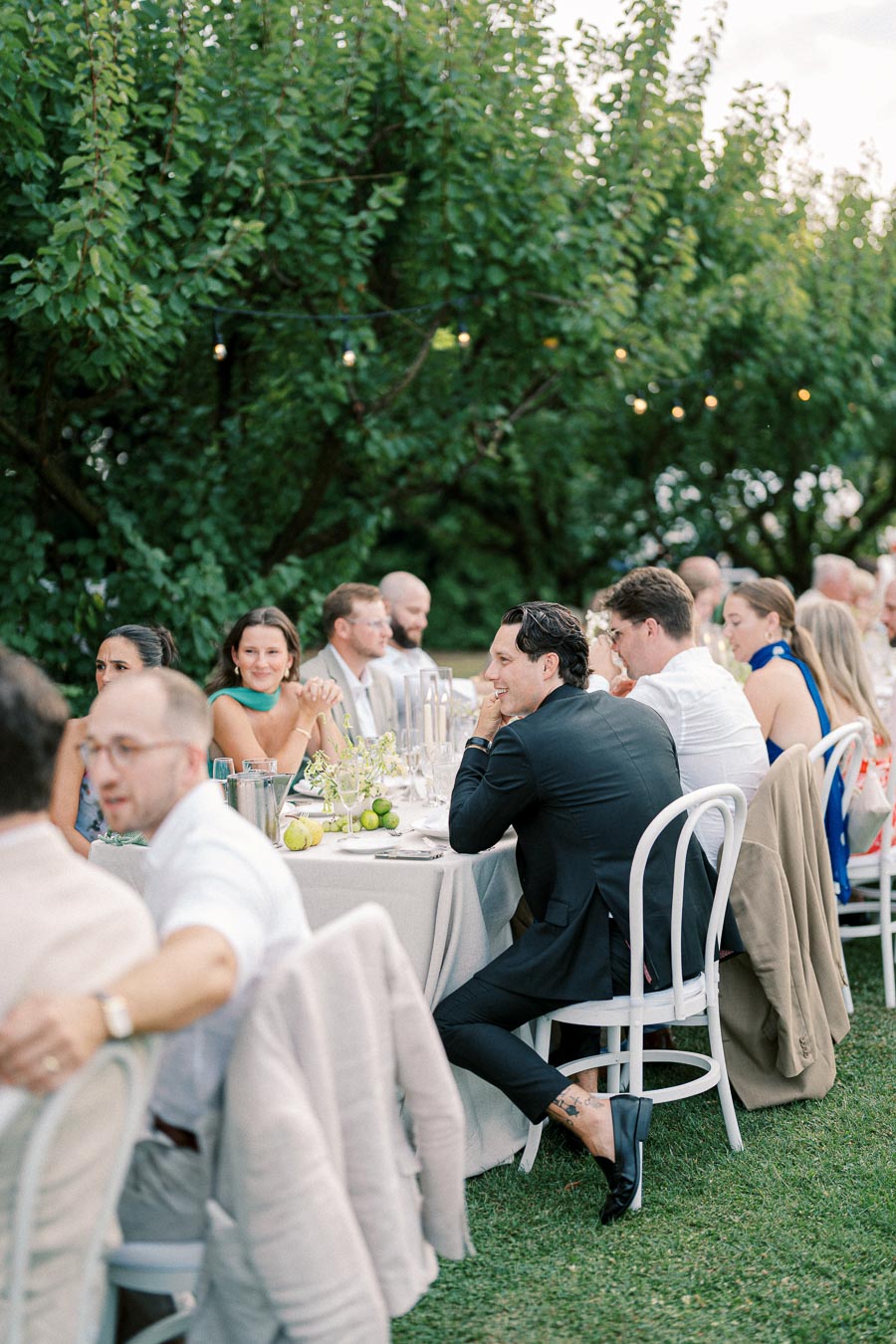 Outdoor garden party with elegantly dressed guests seated at a long table, surrounded by lush greenery and soft string lights, creating a charming and festive atmosphere.