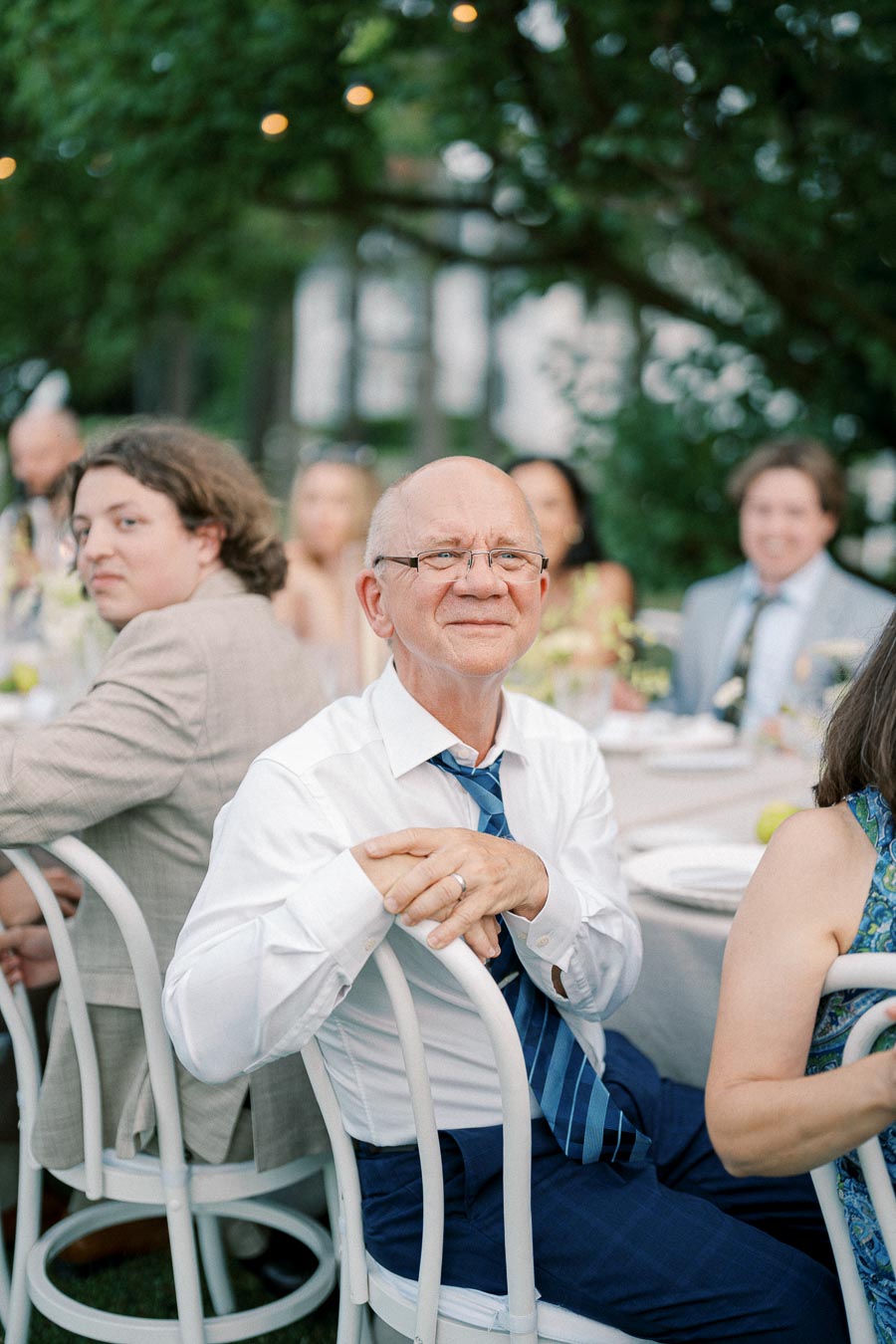 Elderly man smiling at an outdoor gathering, seated at a table with blurred background of other guests, wearing glasses, a white shirt, and a blue tie under a lush green canopy.
