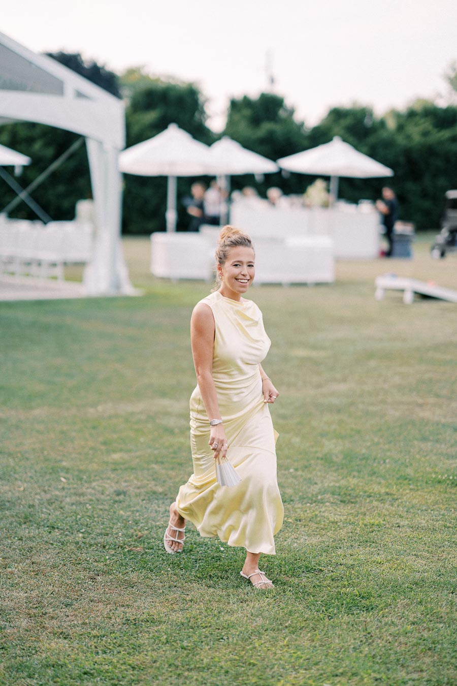 Woman in a light yellow dress smiling and walking on grass at an outdoor event with white tents and umbrellas in the background.
