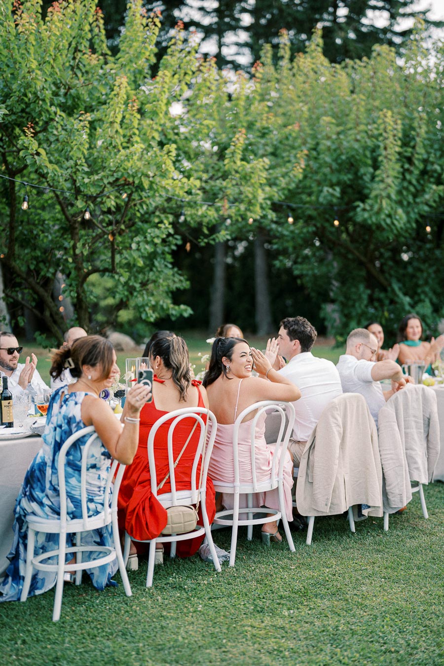 Outdoor garden party setting with elegantly dressed guests seated at a long table, surrounded by lush greenery and decorative lights.