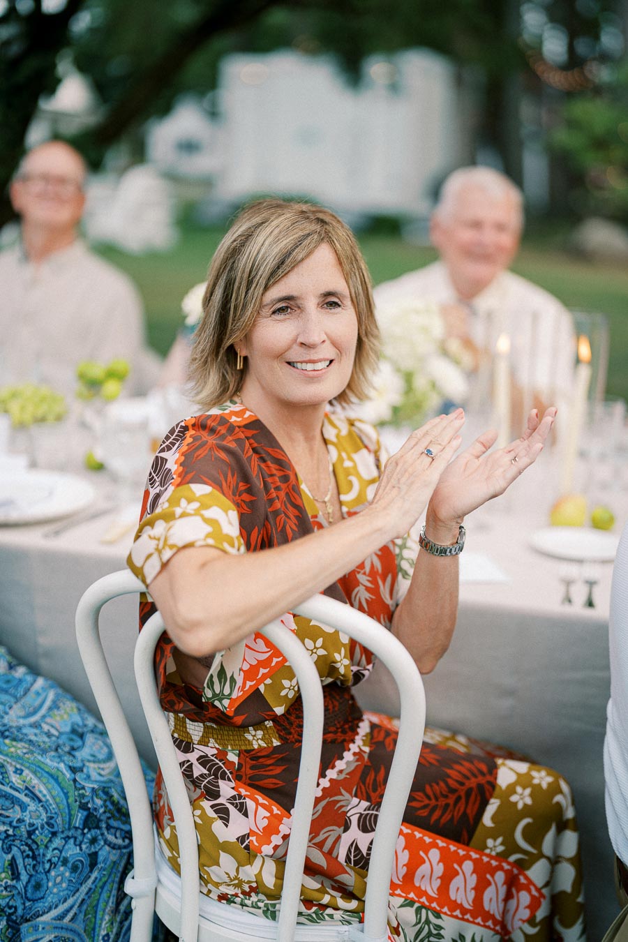 A woman in a colorful floral dress smiling and clapping at an outdoor dinner event, seated at a table set with candles, plates, and greenery.