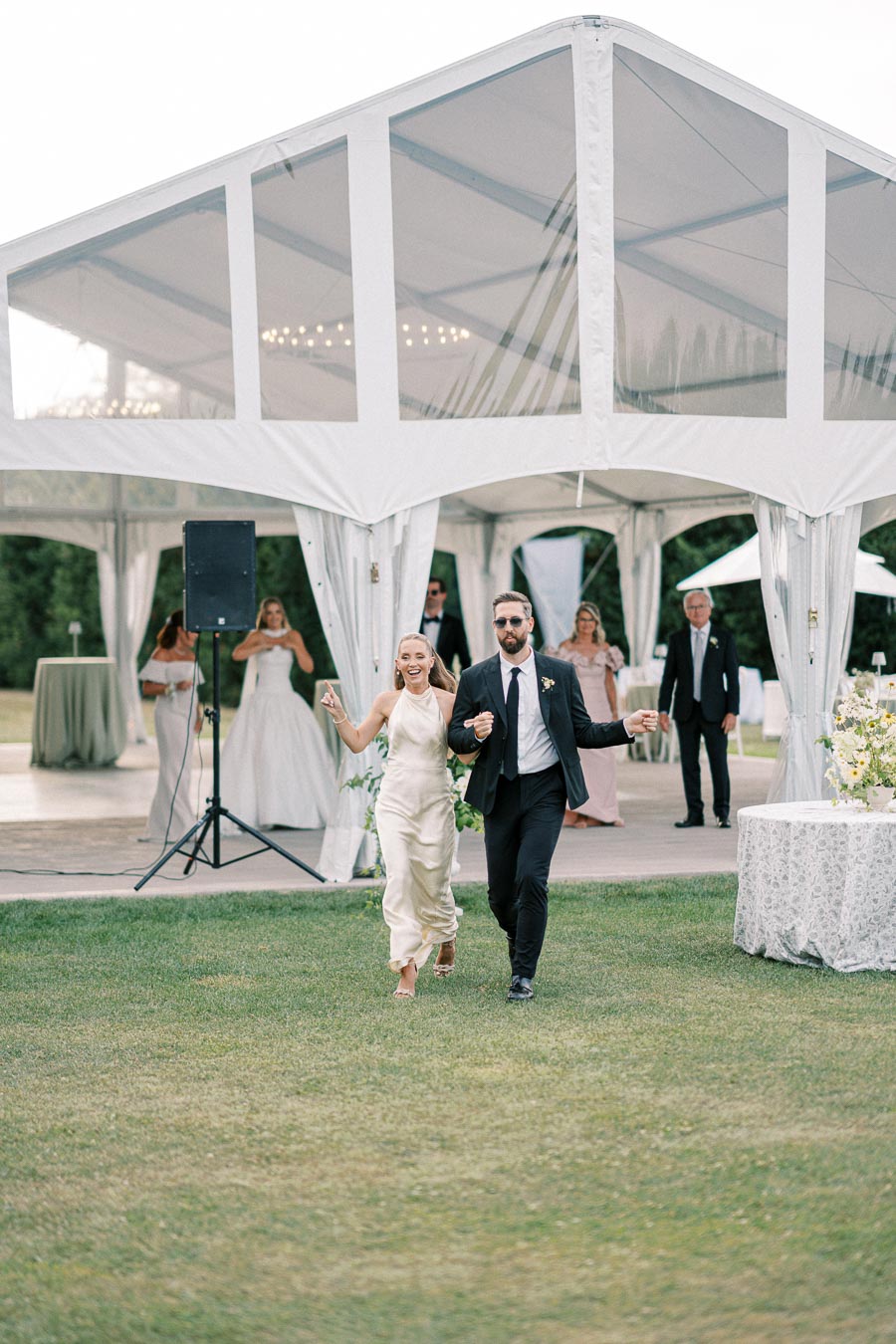 A joyful couple enters a tented outdoor wedding reception, surrounded by elegantly dressed guests and lush greenery.