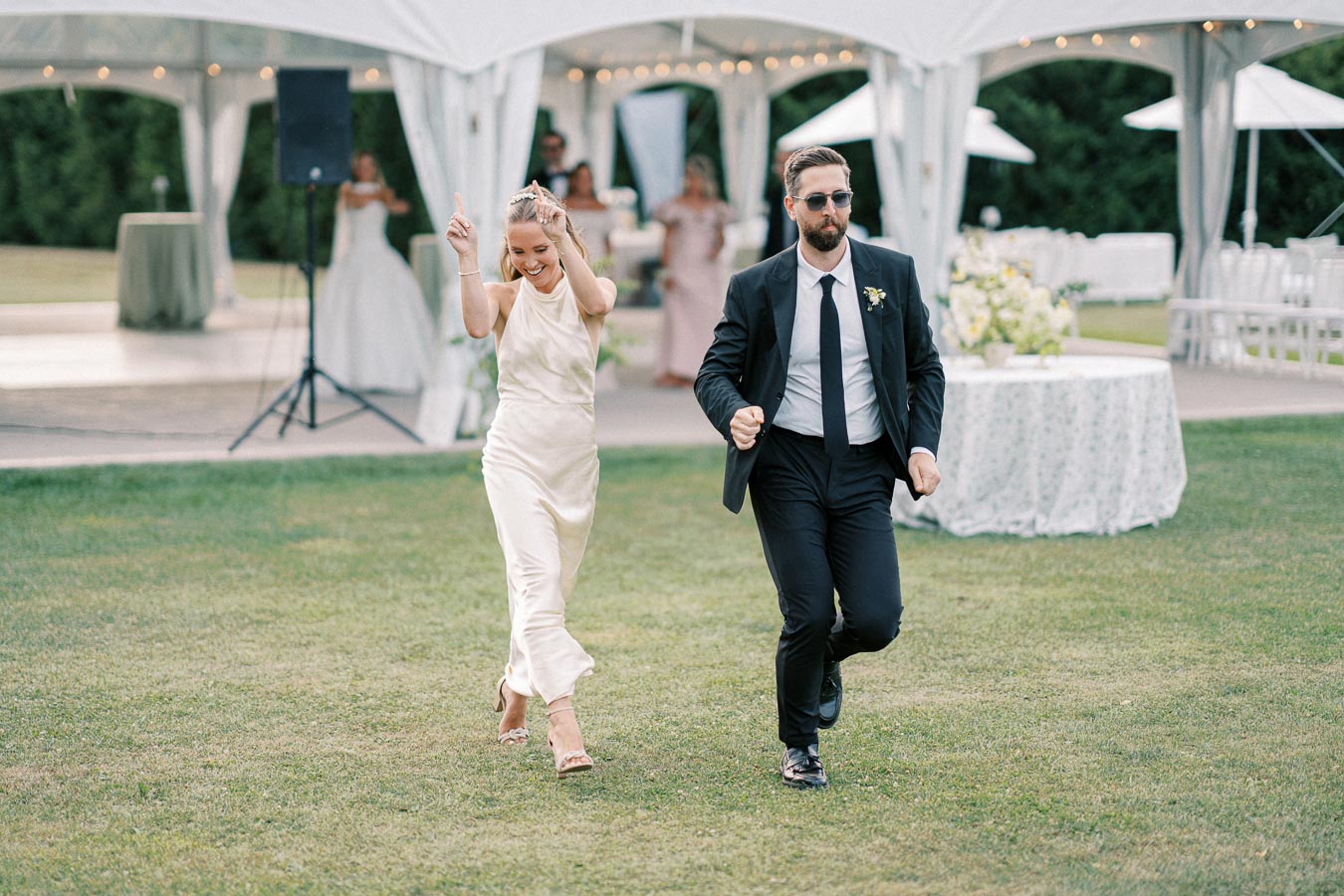 A joyful couple dancing on a grassy lawn at an outdoor wedding reception, the woman wearing a white dress and the man in a suit with sunglasses, surrounded by decorated tables under a white canopy.
