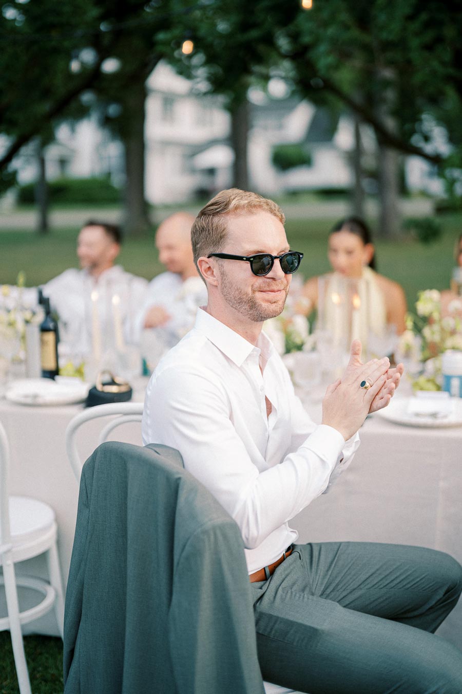 A man in sunglasses and a white shirt claps while seated at an outdoor event, with a jacket draped over his chair and blurred guests in the background.
