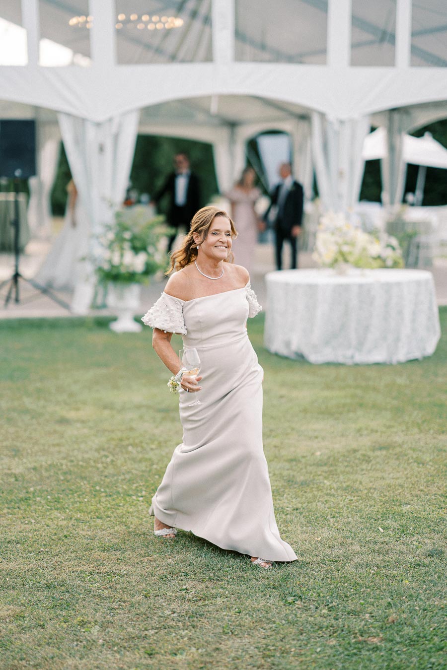 A woman in an elegant off-the-shoulder gown walks on grass, smiling and holding a wine glass at an outdoor event with white tented decor and blurred guests in the background.