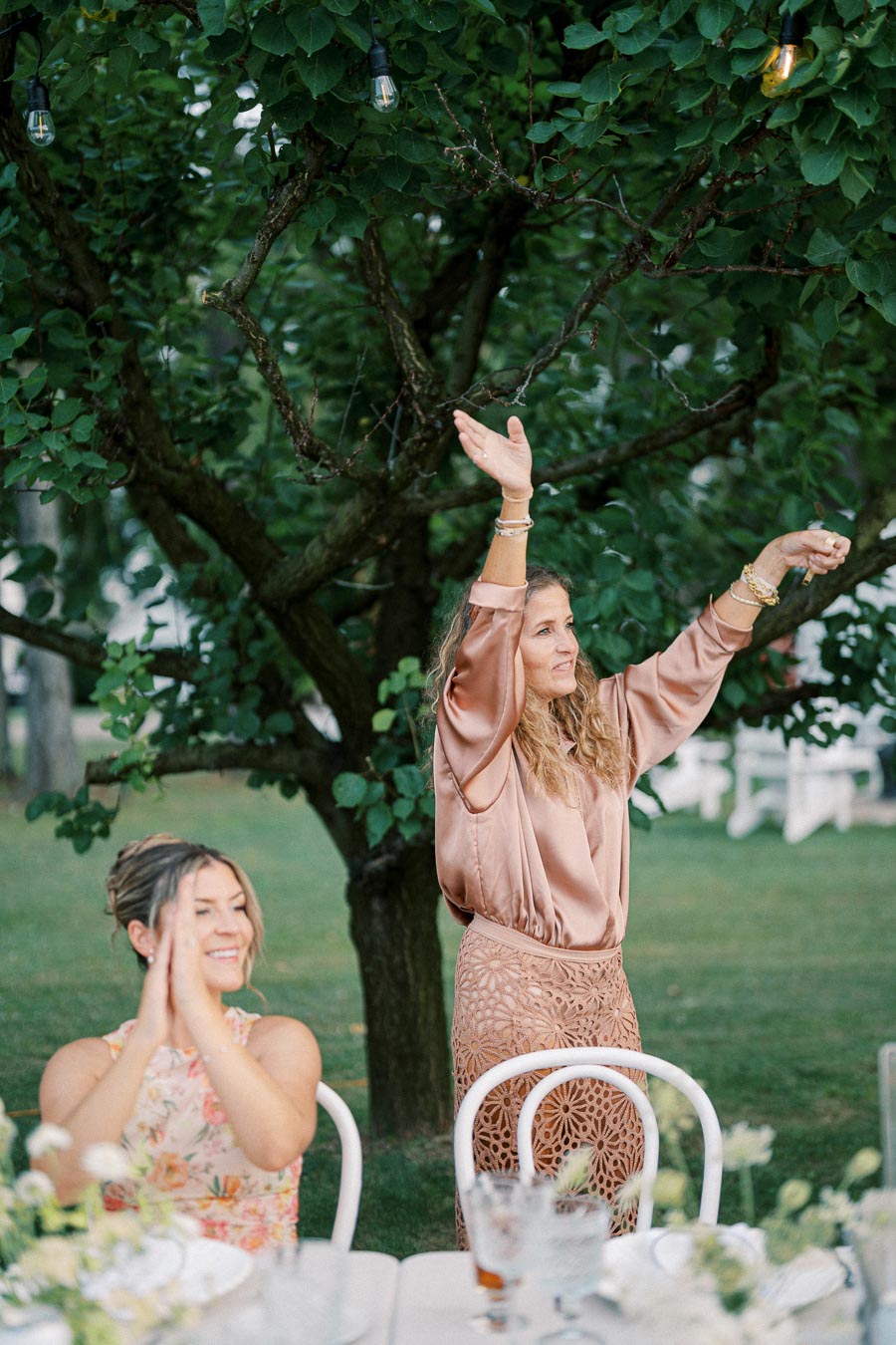 Two women enjoying an outdoor event under a tree, one standing and gesturing with hands raised, and the other sitting and clapping, with tables decorated with flowers and glassware in the foreground.