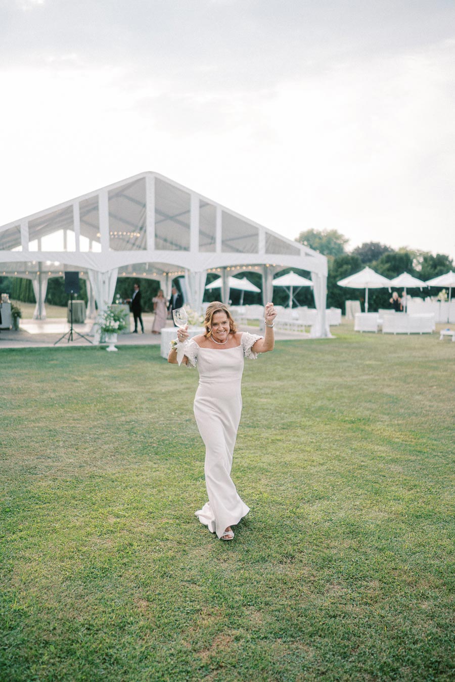 Woman celebrating with a glass of wine in front of a large white event tent on a grassy field, showcasing a joyful outdoor event setting.