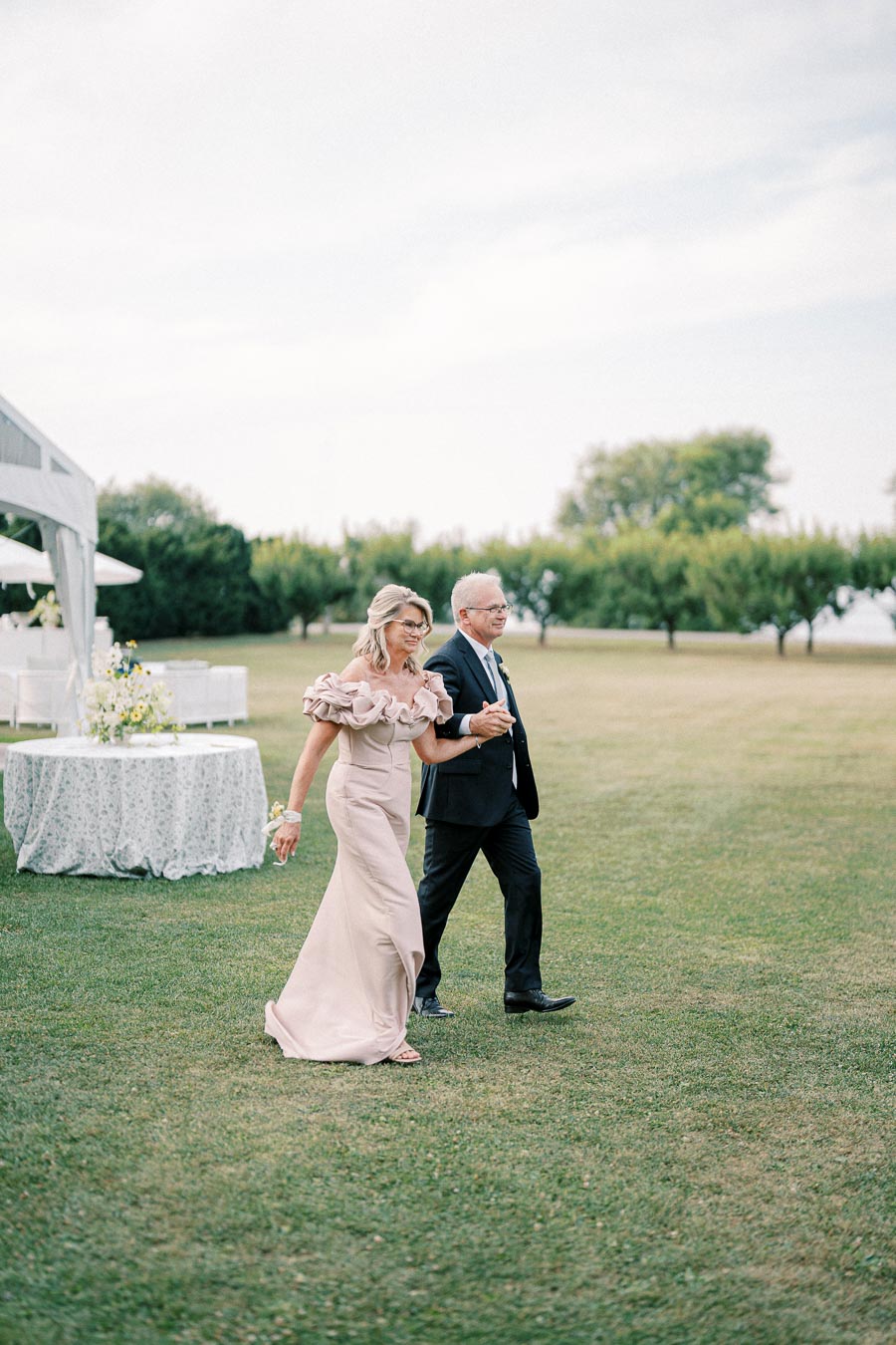 Elegant couple walking on a lush green lawn, with a beautifully decorated canopy visible in the background under a clear sky.