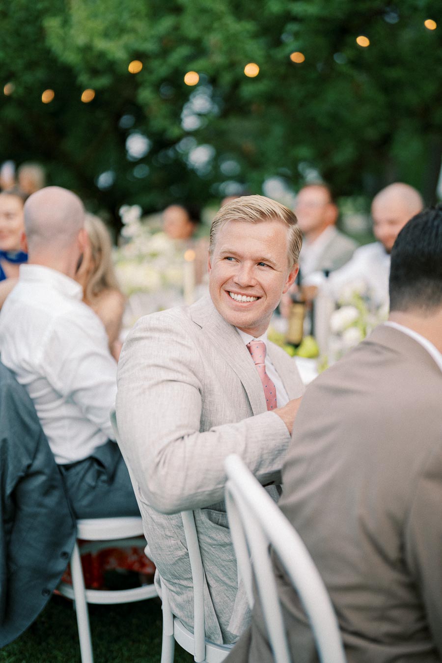Smiling man in a light suit at an outdoor wedding reception with guests seated around decorated tables.
