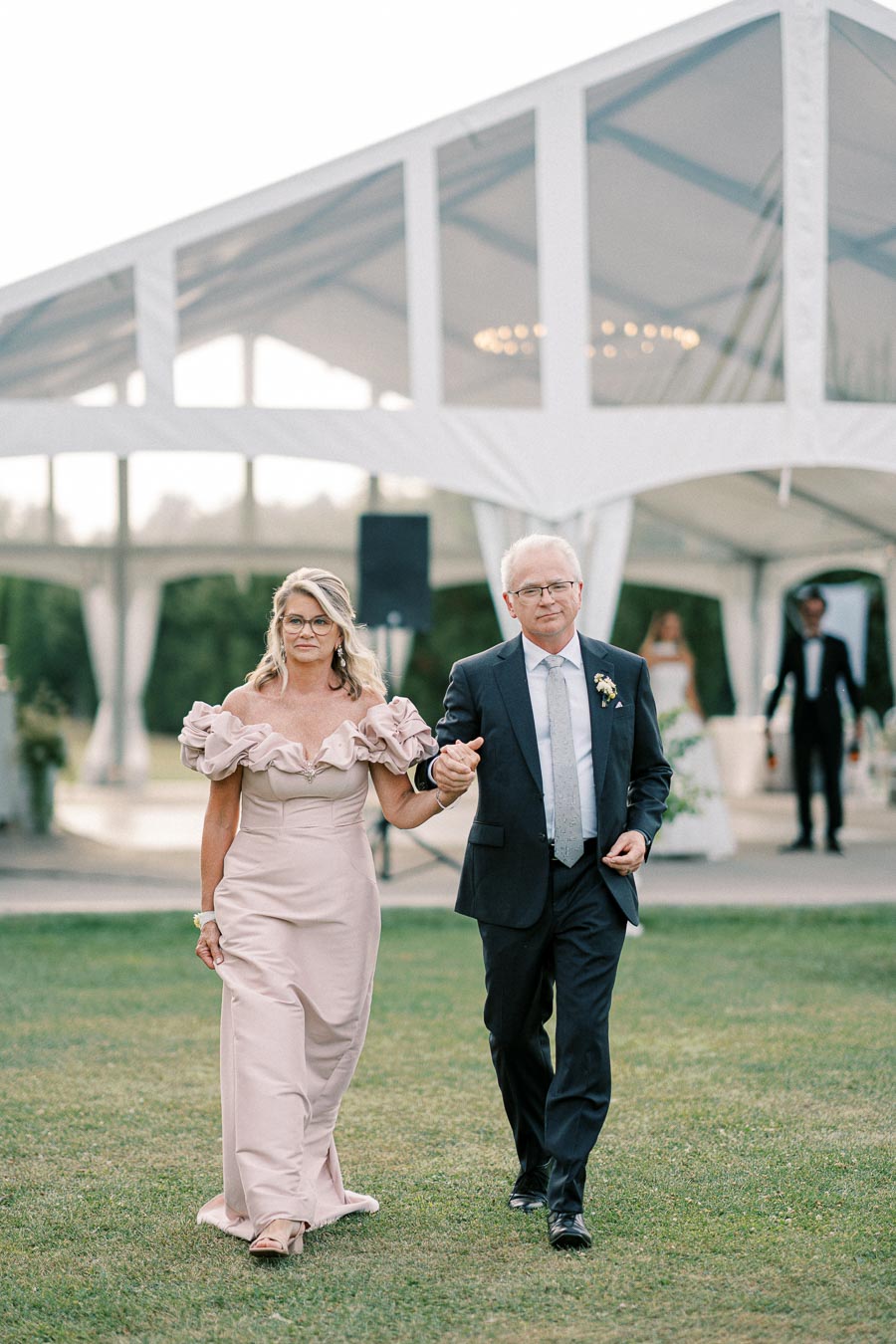 Elegant couple walking hand in hand at an outdoor event, with a sophisticated tent structure in the background. The woman is wearing a pink off-shoulder dress, and the man is dressed in a formal suit with a boutonnière.