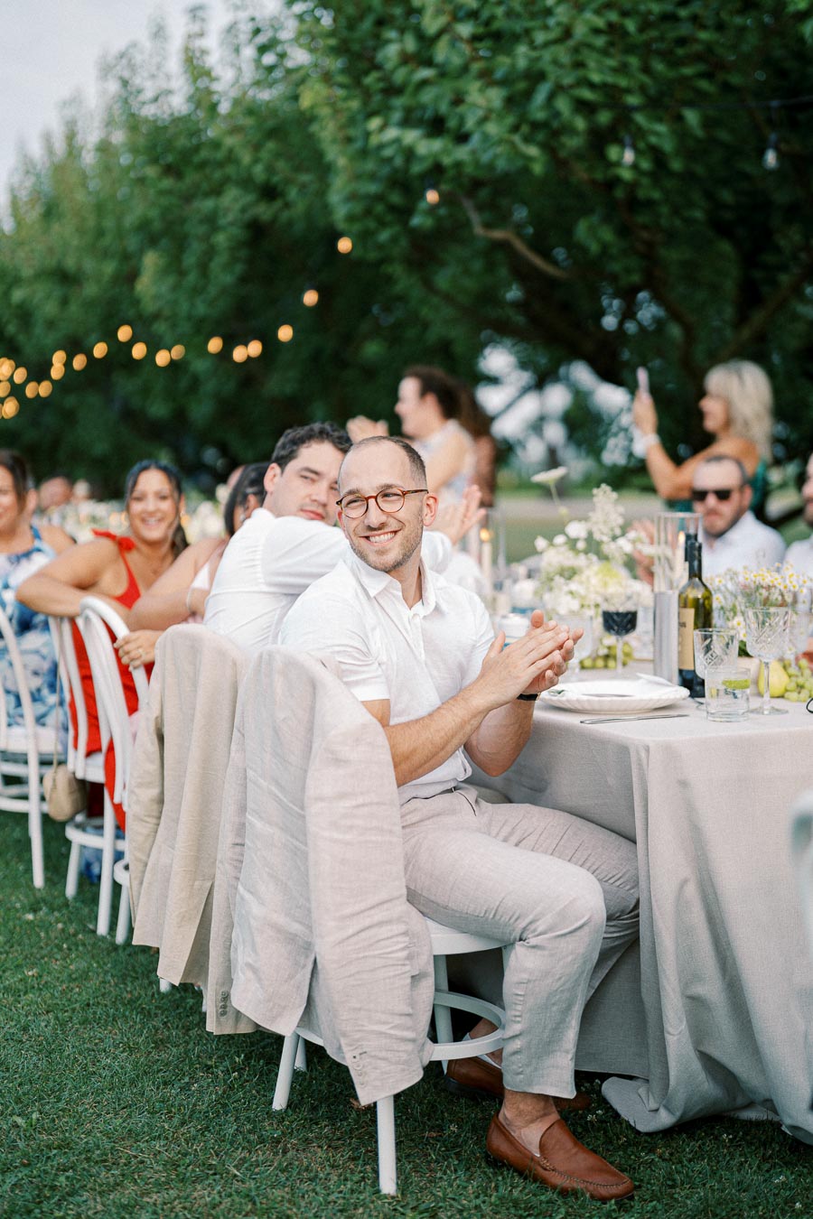 A joyful outdoor wedding reception with guests seated at a long table, decorated with elegant floral arrangements and string lights. A man in a white shirt and glasses smiles while clapping, creating a warm and festive atmosphere.