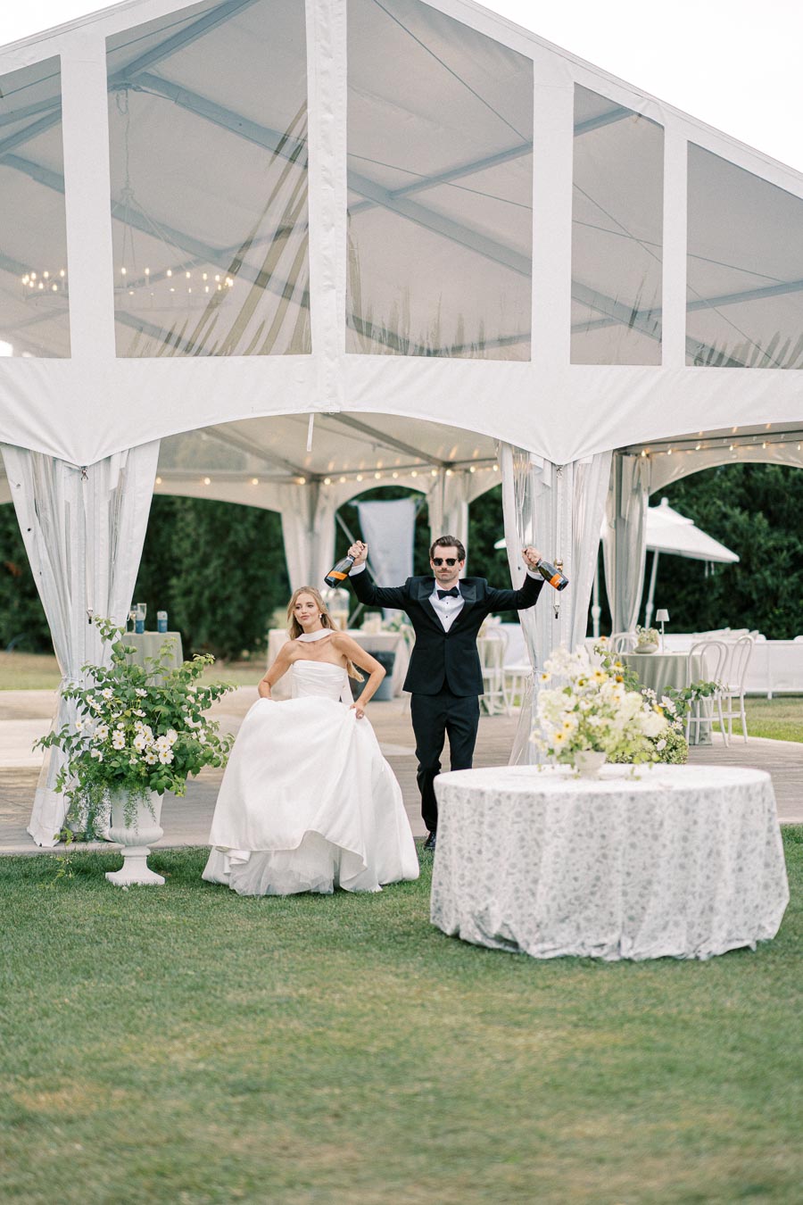 Elegant outdoor wedding reception with bride in stylish white gown and groom in black tuxedo celebrating with champagne bottles, under a beautifully decorated white tent, surrounded by floral arrangements and green lawns.