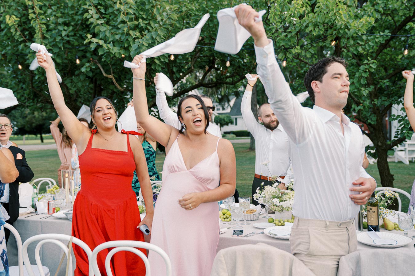 Group of people celebrating outdoors at a wedding reception, waving napkins joyfully in the air.