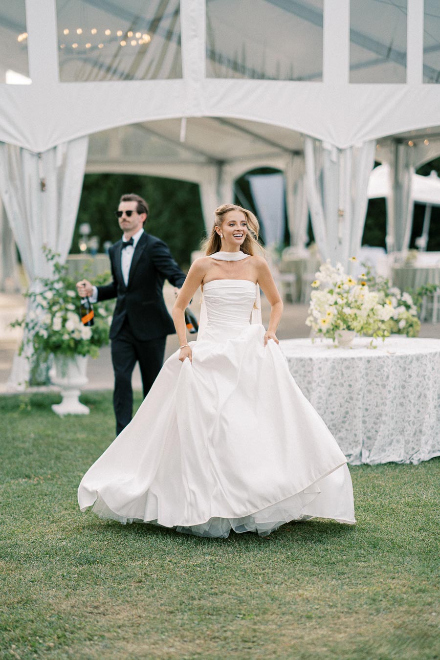 A joyful bride wearing a stunning white wedding dress walks on the grass at an outdoor celebration, with a groom in a tuxedo holding champagne bottles in the background, under a beautifully decorated tent.