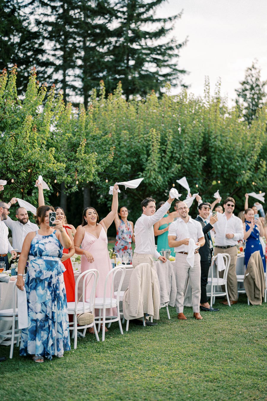 Group of people celebrating outdoors at an elegant garden party, wearing formal attire and waving napkins, with lush greenery and trees in the background.