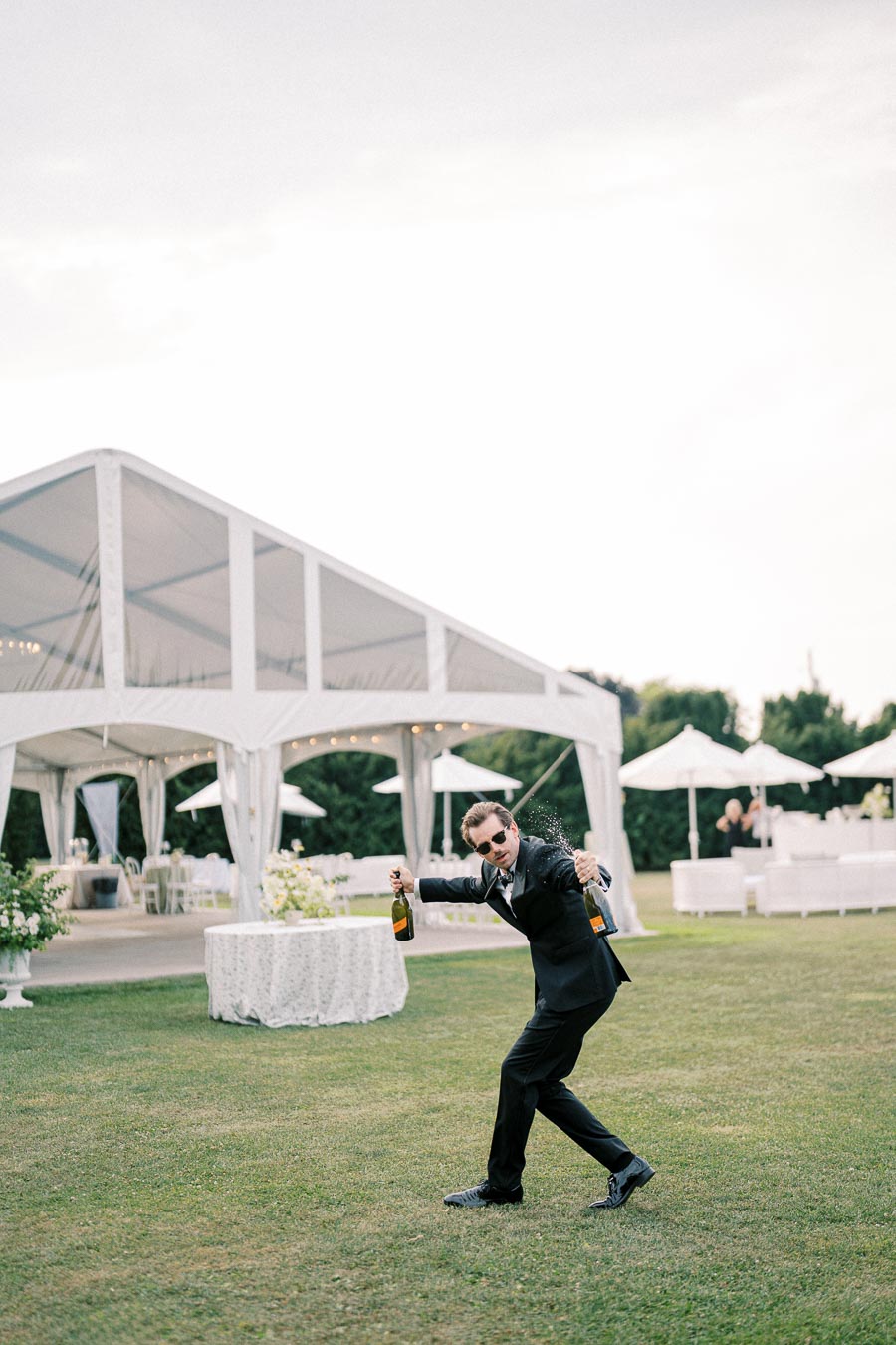 A man in a black suit and sunglasses playfully sprays champagne from two bottles in an outdoor wedding reception setting, with a white tent and elegant tables in the background.