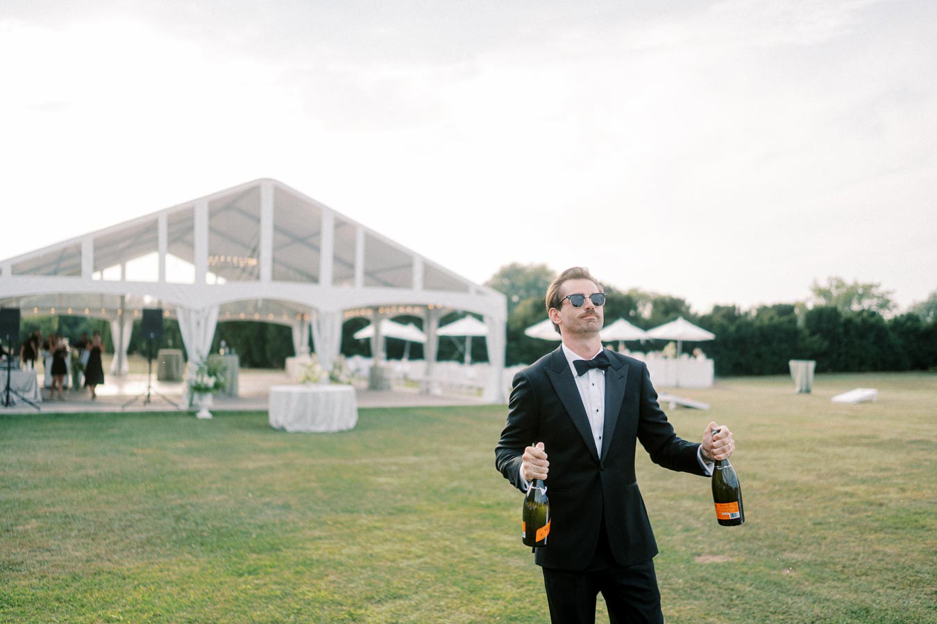Man in sunglasses and tuxedo holding champagne bottles outdoors at an elegant event with a white marquee tent in the background.