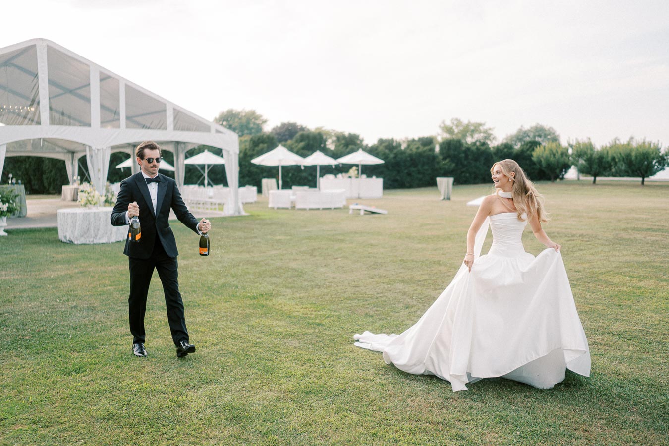 Elegant outdoor wedding scene with a bride in a white gown and groom in a black tuxedo holding champagne bottles, set against a backdrop of a white marquee and lush greenery.