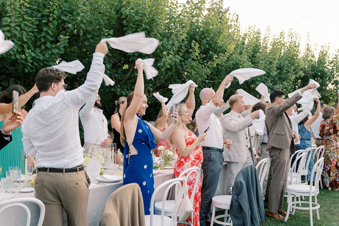 People at an outdoor event waving white napkins in celebration around a long dining table set with decorative elements.