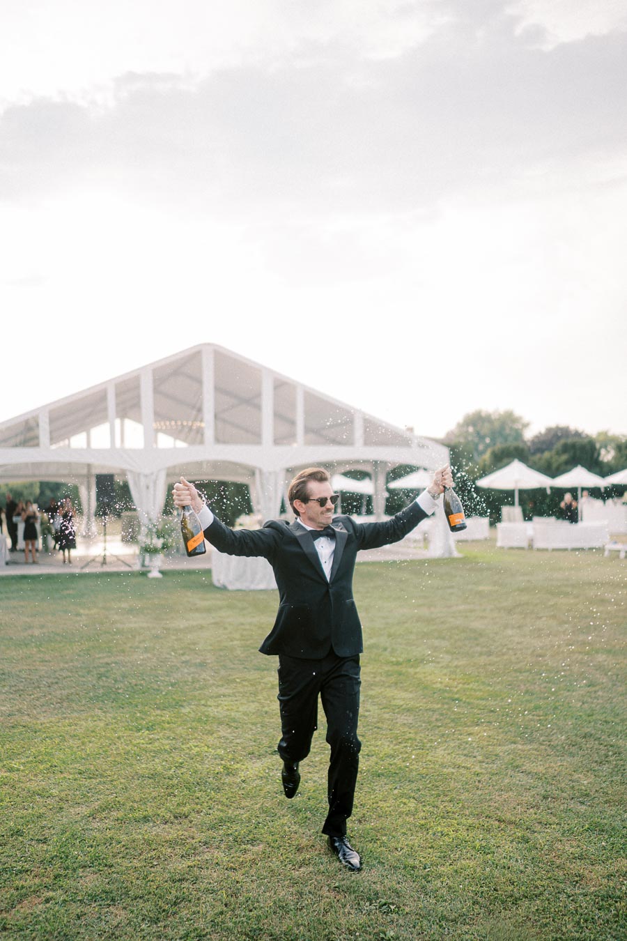 Man in a tuxedo joyfully running with champagne bottles at an outdoor event, spraying champagne near a large white tent.