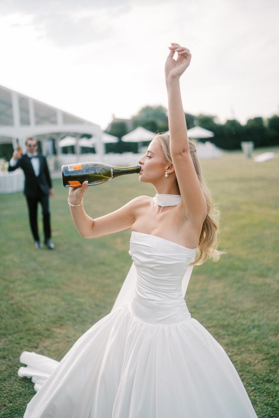 Bride joyfully drinking champagne from a bottle in a sunny outdoor setting, wearing a white wedding dress, with a man in a suit holding a drink in the background.