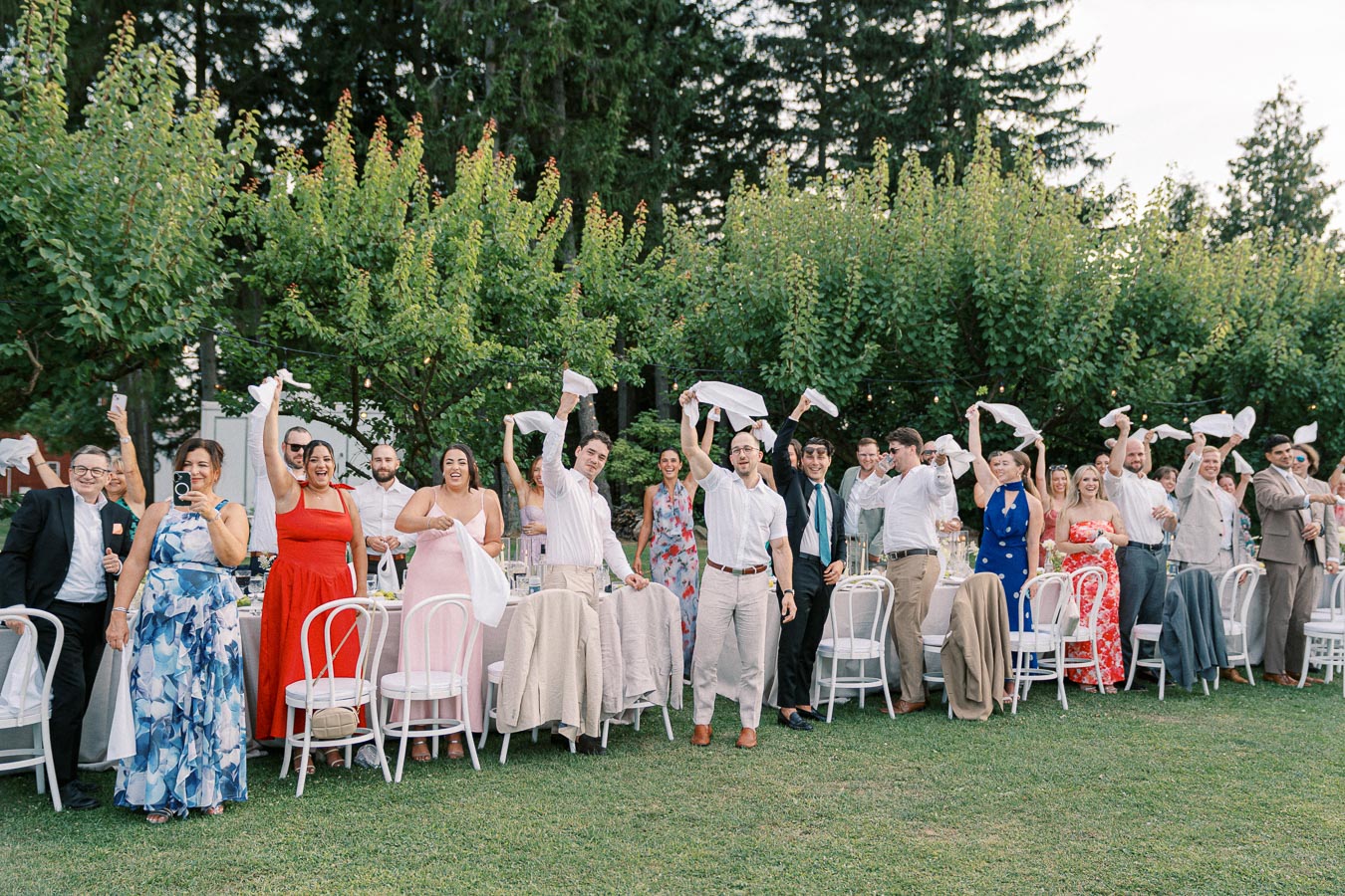 Group of happy wedding guests celebrating outdoors, waving napkins, with trees in the background.