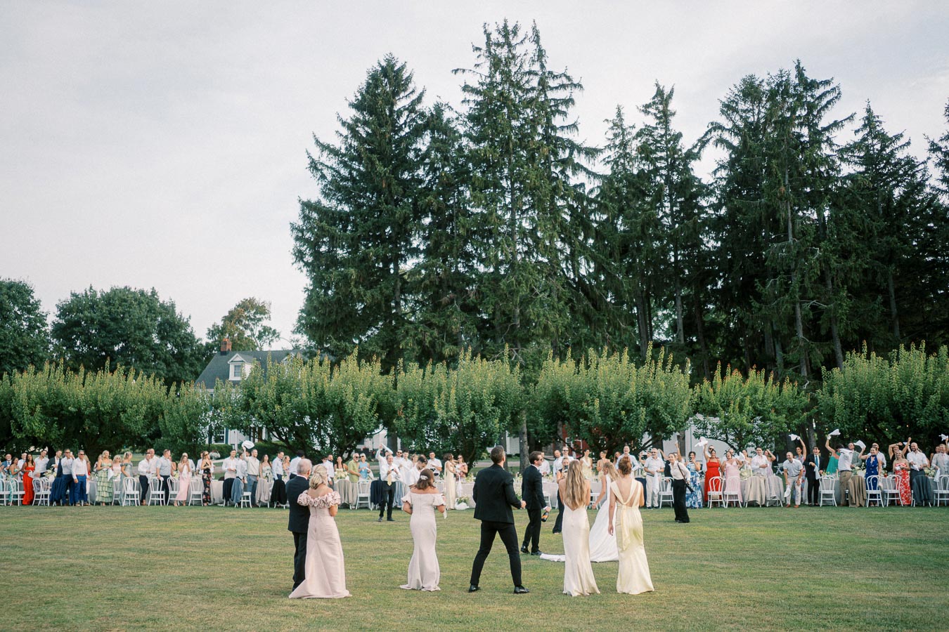 Elegant outdoor wedding reception with guests gathered on a lush green lawn, surrounded by tall trees, celebrating a newlywed couple under a clear sky.