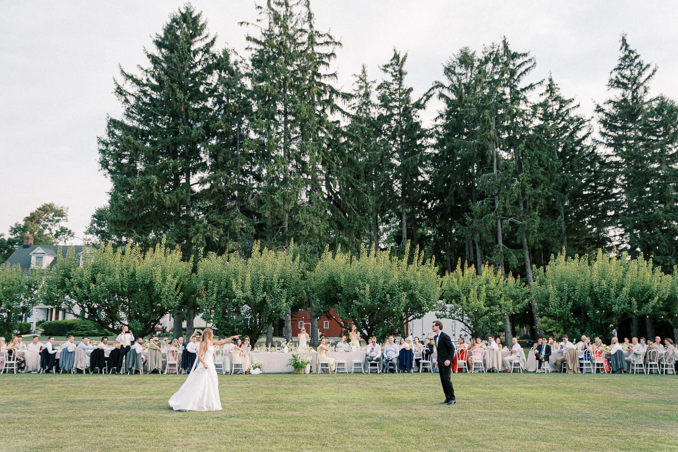 Outdoor wedding reception featuring a bride and groom dancing on a lush green lawn, surrounded by guests seated at elegantly decorated tables, with a scenic backdrop of tall trees.