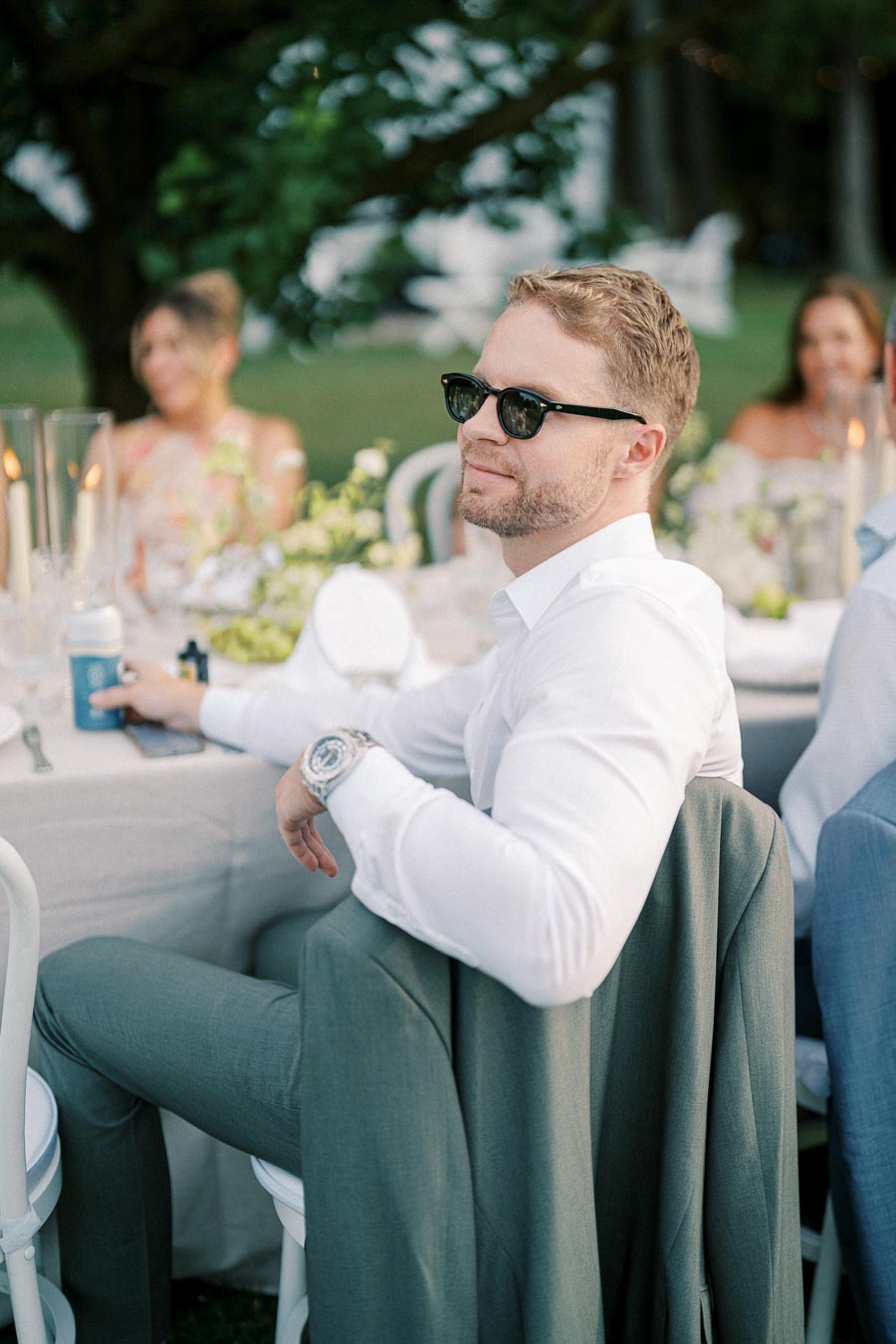 Man in sunglasses sitting at an elegant outdoor event, wearing a white shirt and gray suit jacket draped over the chair, with lush green surroundings and a beautifully set table in the background.
