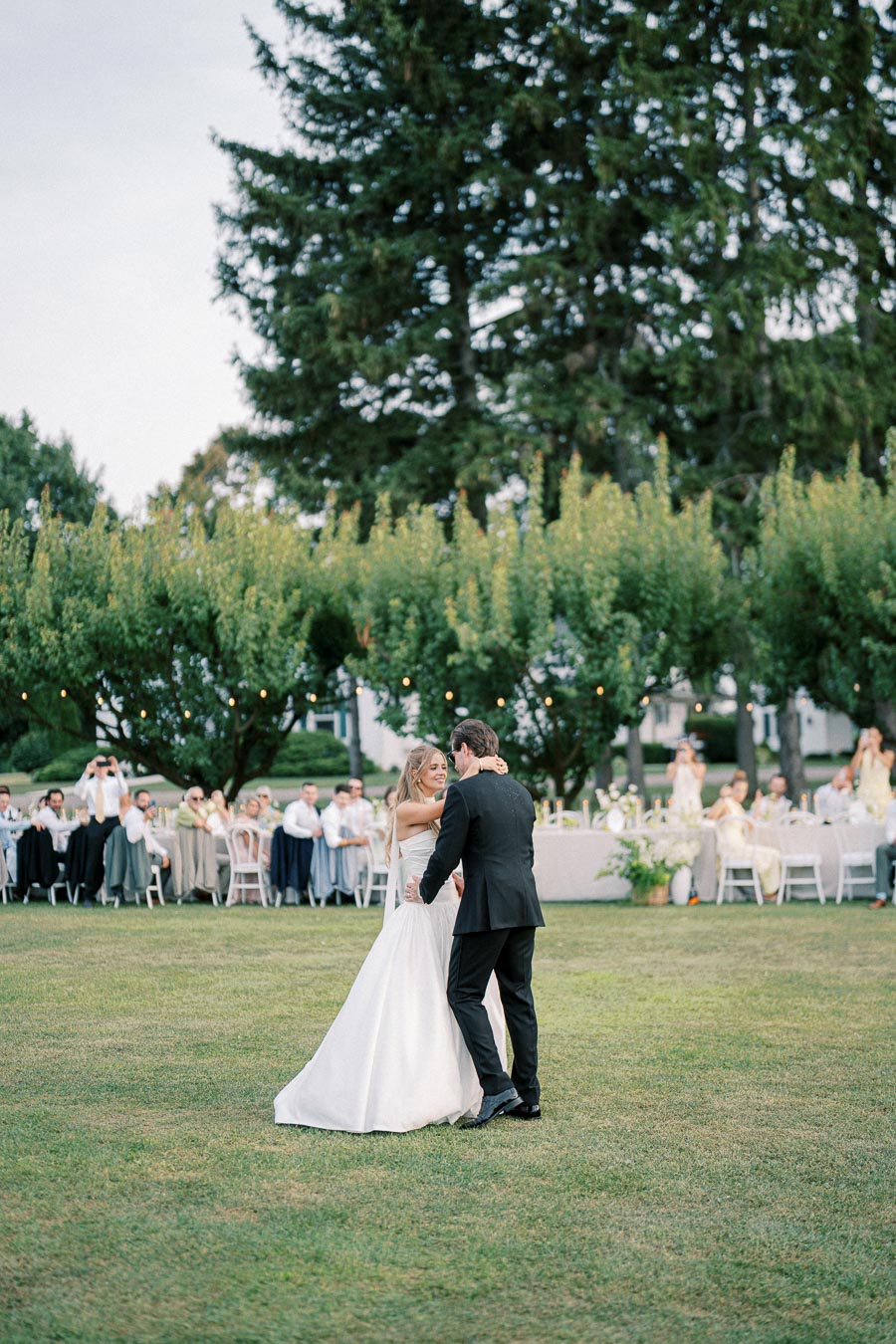 A bride and groom sharing a first dance on a grassy lawn at an outdoor wedding, surrounded by guests seated at decorated tables under trees with string lights.