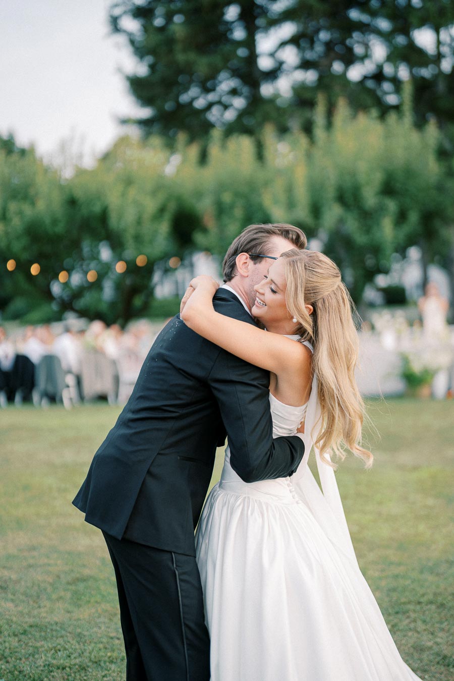 Newlywed couple embracing joyfully outdoors in a lush garden setting during their wedding celebration, with the bride wearing an elegant white dress and the groom in a classic black suit.