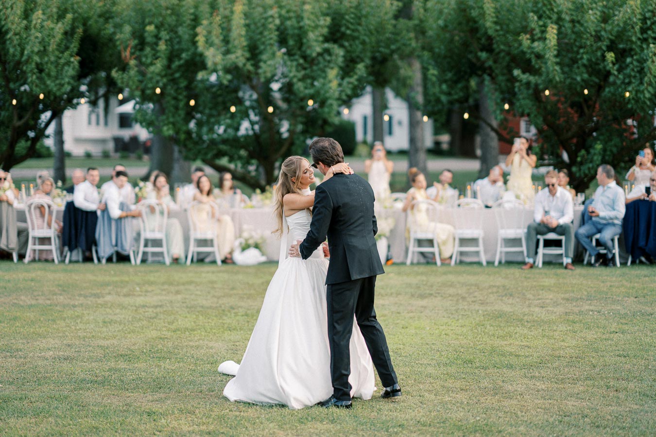 Newlywed couple sharing their first dance outdoors at a garden wedding reception, surrounded by seated guests under string lights.