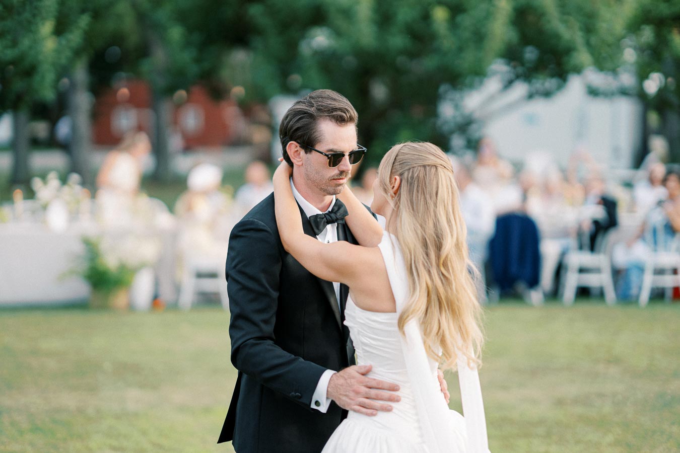 A bride and groom share a dance outdoors at their wedding, surrounded by seated guests and lush greenery. The bride has long blonde hair and wears a white gown, while the groom is in a black tuxedo and sunglasses.